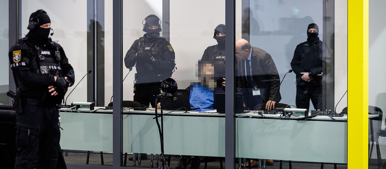 Ein Angeklagter sitzt von Polizisten bewacht hinter einer Glasscheibe.