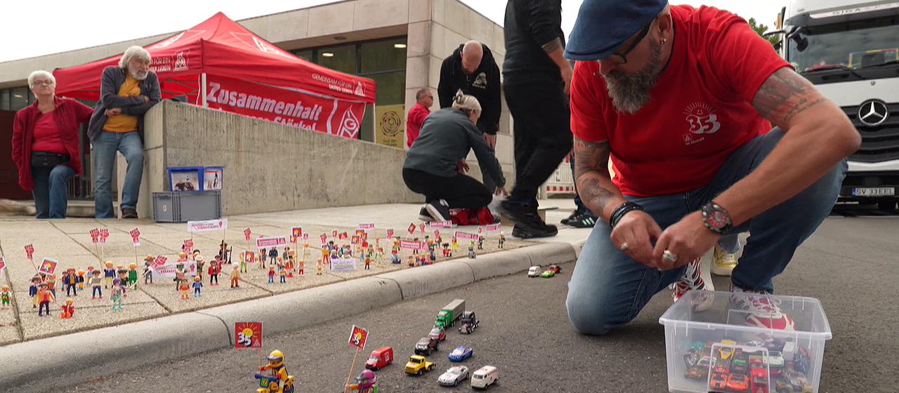 Menschen bauen vor einem Stand der IG-Metall Spielzeugautos auf einer Fahrbahn auf.