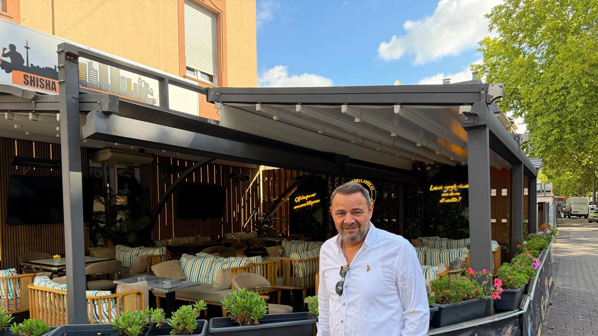 Ein mittelalter Mann im hellen Hemd und in grüner Hose steht vor der teils überdachten Terrasse eines Lokals. Er lächelt in die Kamera.  | hessenschau.de