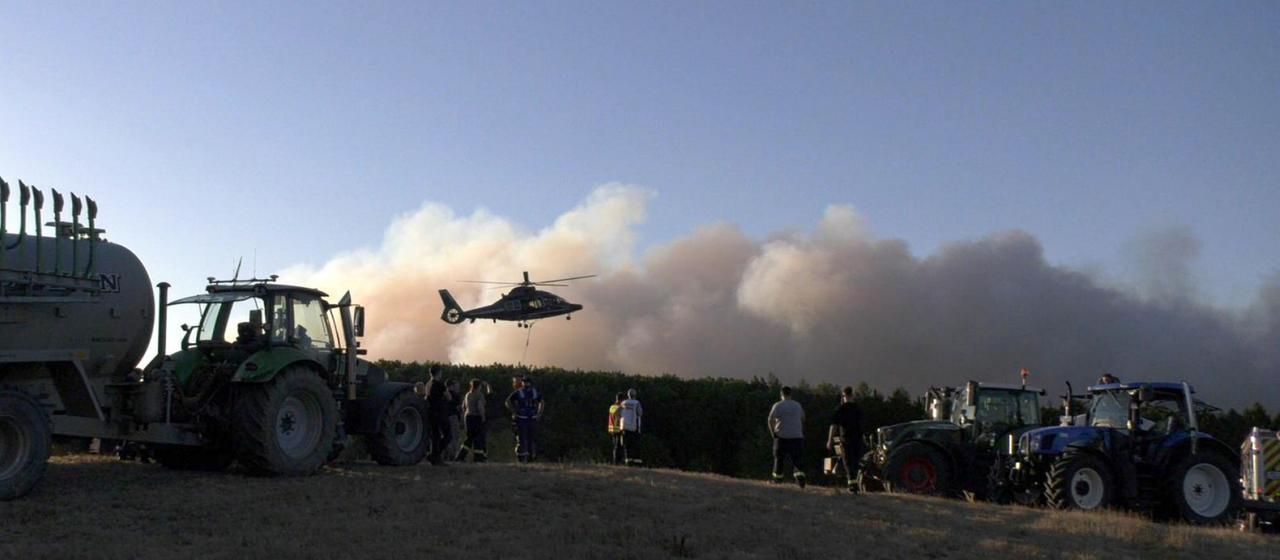 Ein Traktor und Menschen stehen nahe eines Waldes in der Eifel, über dem Rauch eines Waldbrandes aufsteigt. Ein Helikopter startet in diesem Moment.