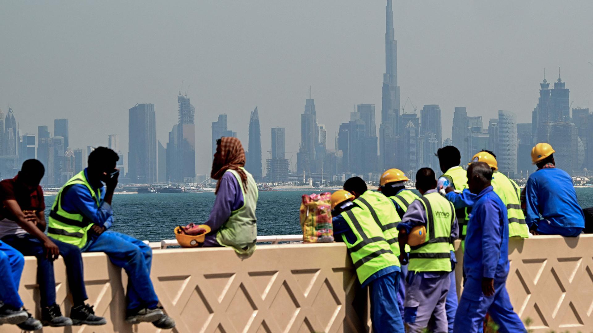 Arbeiter sitzen vor der Skyline der Stadt Dubai auf einer Mauer. | AFP