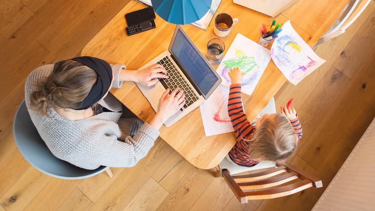 Ein zweijähriges Kind malt ein Bild, während seine Mutter Zuhause im Homeoffice an einem Laptop arbeitet.