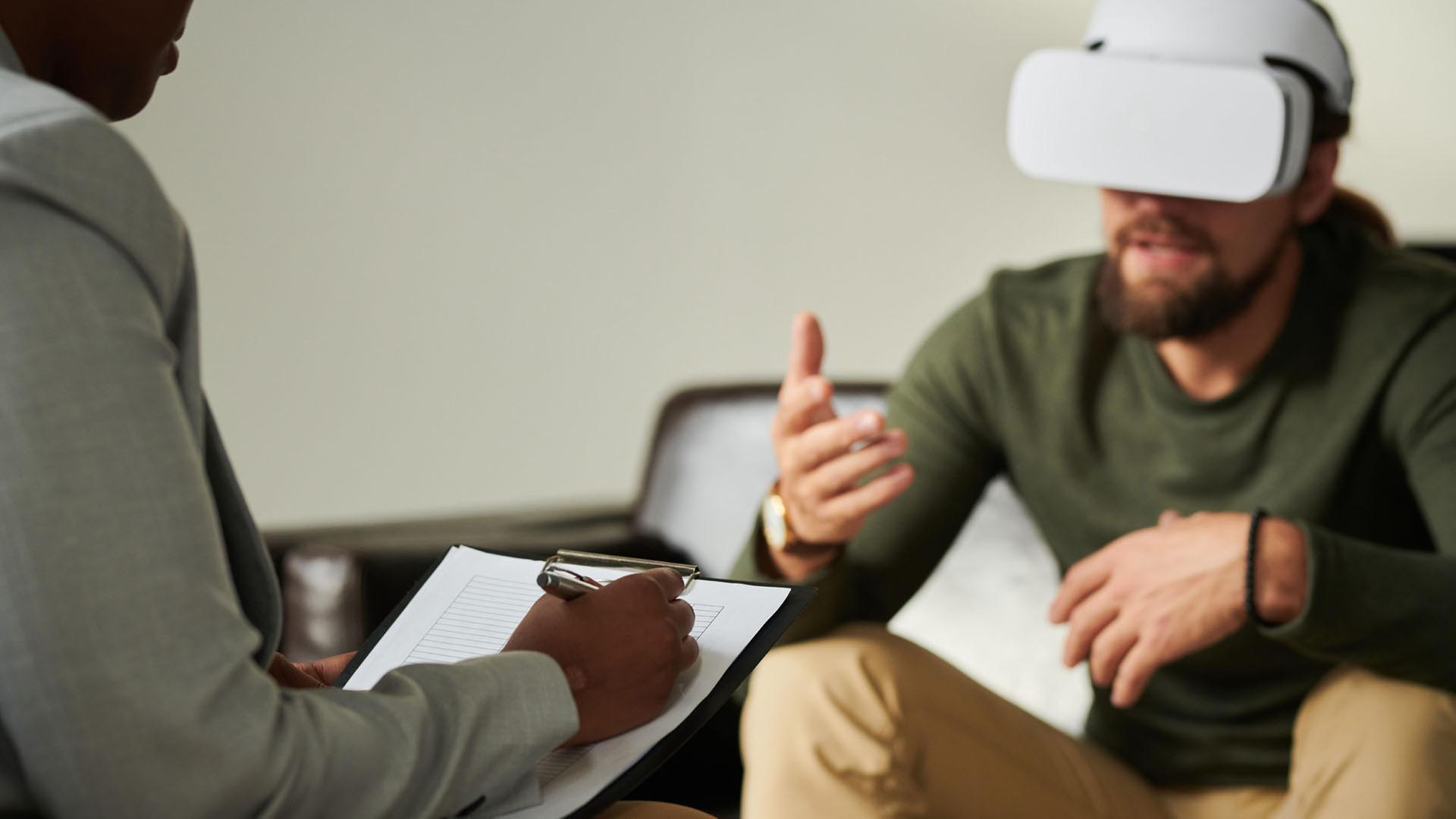Eine Person mit einer VR-Brille sitzt einer anderen Person mit Klemmbrett gegenüber. | picture alliance / AnnaStills