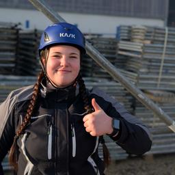 Foto: Eine junge Frau steht vor einem Stapel Stahl-Gerüstteile. Sie trägt eine schwarze Arbeitsjacke und einen blauen Helm. Mit ihrer linken Hand zeigt sie Daumen hoch. 