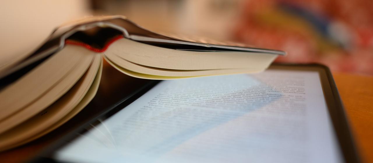 Ein iPad Tablet mit einer geöffneten eBook und ein Buch liegen auf einem Tisch.