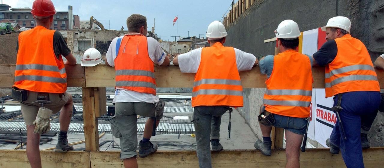Bauarbeiter stehen in ihrer Pause an einem Zaun vor einer Baugrube.