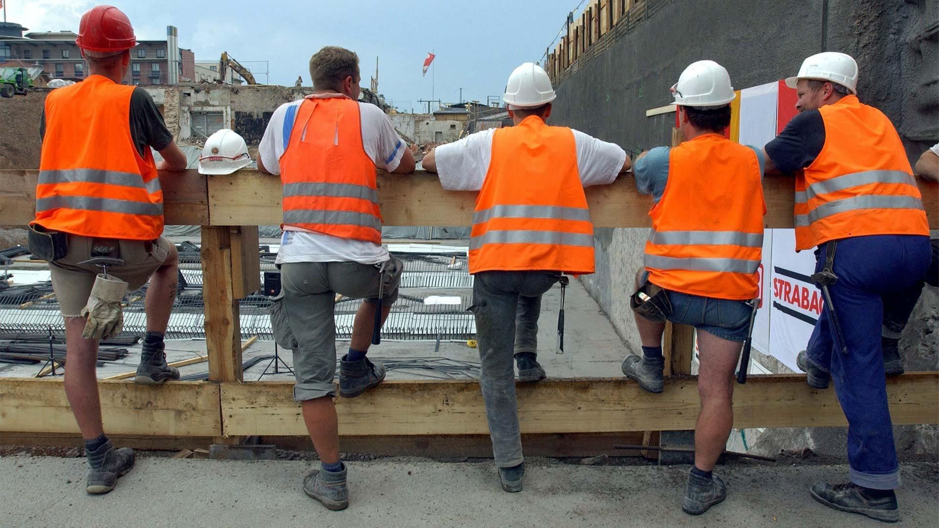 Bauarbeiter stehen in ihrer Pause an einem Zaun vor einer Baugrube. | picture-alliance / ZB