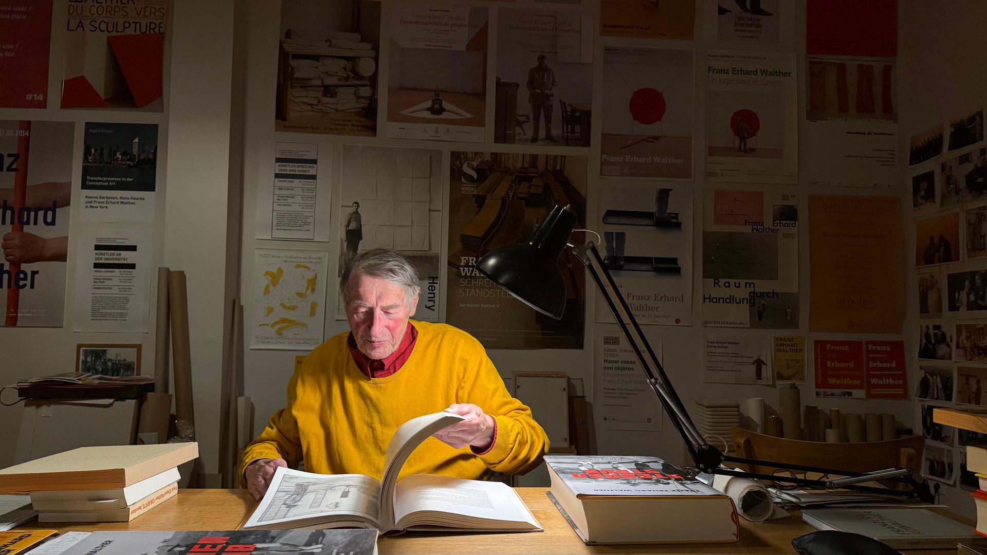 Ein Mann mit grauem Haar, rotem Hemd und gelbem Pullover darüber sitzt an einem großen Tisch in der Dunkelheit nur mit einer Tischlampe beleuchtet und blättert in einem großen Buch. Im Hintergrund an der Wand sind im schummrigen Licht viele Plakate zu sehen. | Paul Mosbacher (hr)
