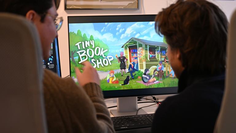David Zapfe-Wildemann und Raven Rusch sitzen vor einem Bildschirm, auf dem die Startseite des Spiels "Tiny Bookshop" zu sehen ist.