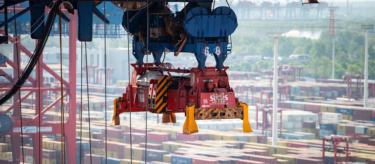 Ein leerer Greifer einer Containerbrücke schwebt über einem Schiff. 
