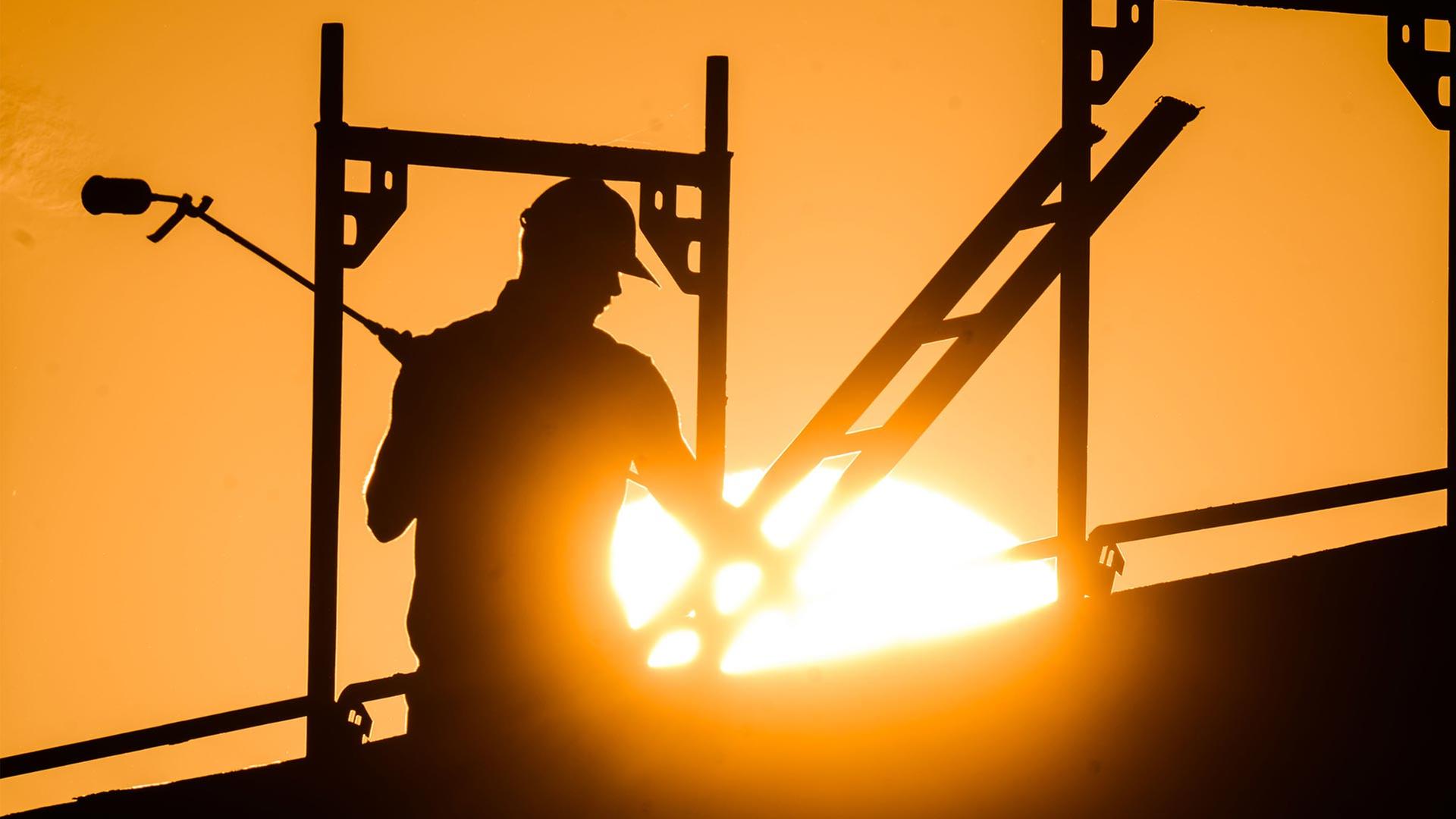 Bauarbeiter mit Gasbrenner auf einem Baugerüst, Sonne im Hintergrund. | picture alliance/dpa