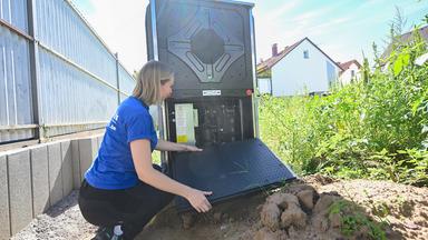 Eine Mitarbeiterin in einem Handwerksbetrieb für Heizungsbau, inspiziert vor einem neu gebauten Wohngebäude im Garten eine Wärmepumpe.