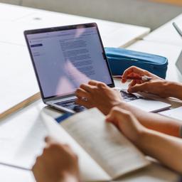 Schüler sitzen mit einem Buch, Laptop und Tablet an einem Tisch.