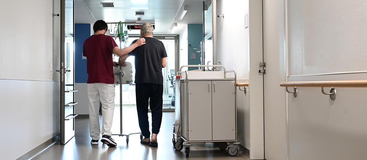 Ein Pfleger geht mit einem Patienten über einen Krankenhausflur. 