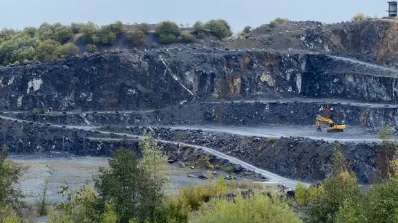 Ein Bagger steht vor dem dunklen Fels des Steinbruchs.