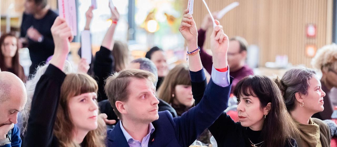 Kerstin Wolter, Maximilian Schirmer und Elif Eralp sitzen nebeneinander in einem Saal und heben Abstimmungskarten in die Luft.