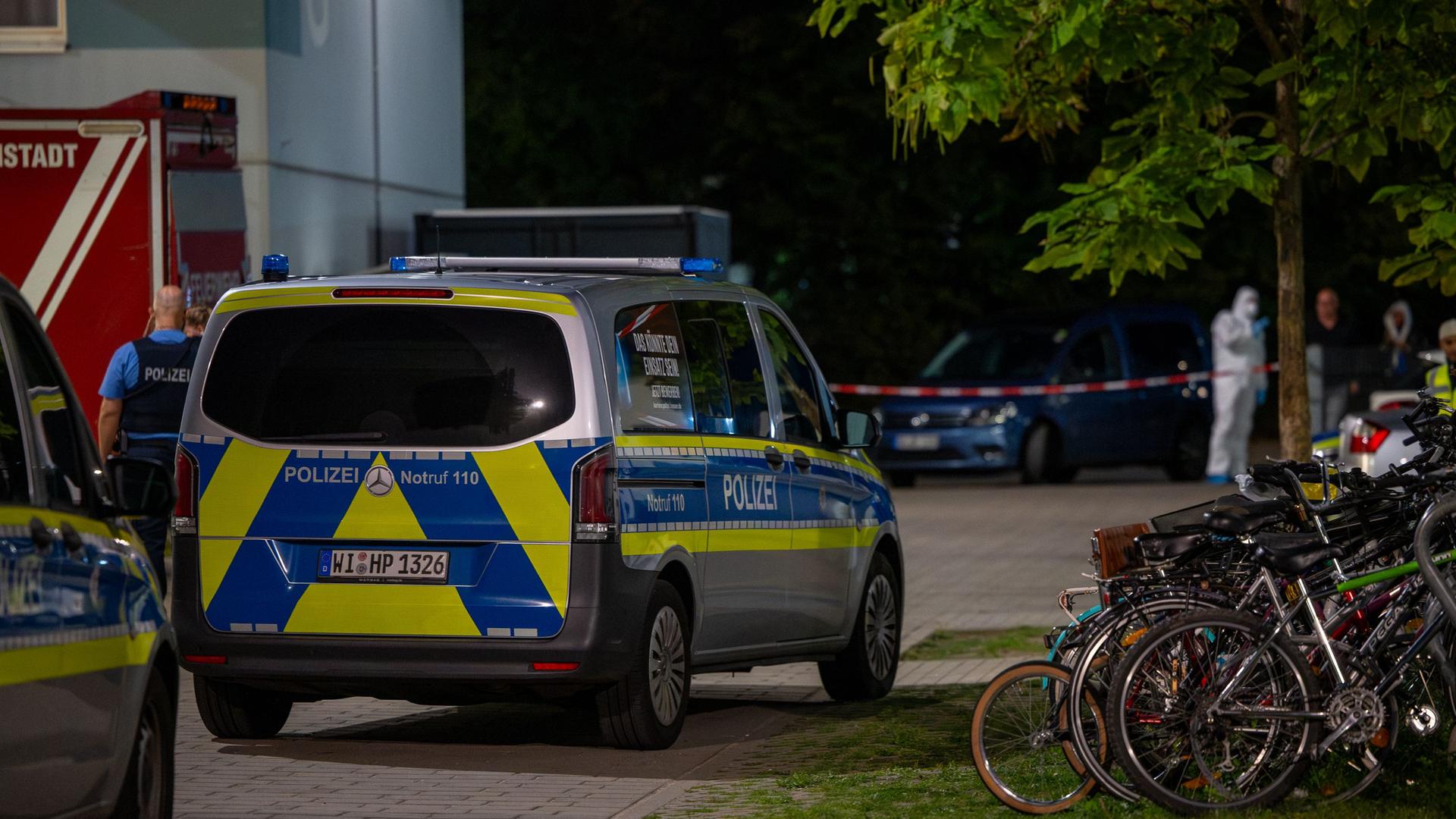 Ein Polizeiauto steht auf einem Gelände neben Fahrradständern und einem Baum. Im Hintergrund sind verschwommen Einsatzkräfte in weißem Schutzanzug zu erkennen. | 5vision.news