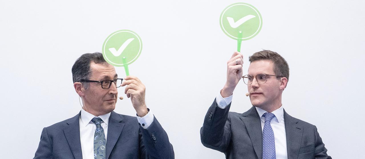 Cem Özdemir und Manuel Hagel halten grüne Schilder mit einem weißen Haken darauf in der Hand.