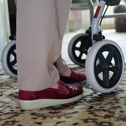 Füße einer Frau gehen hinter den Rädern eines Rollators in einer Wohnung.