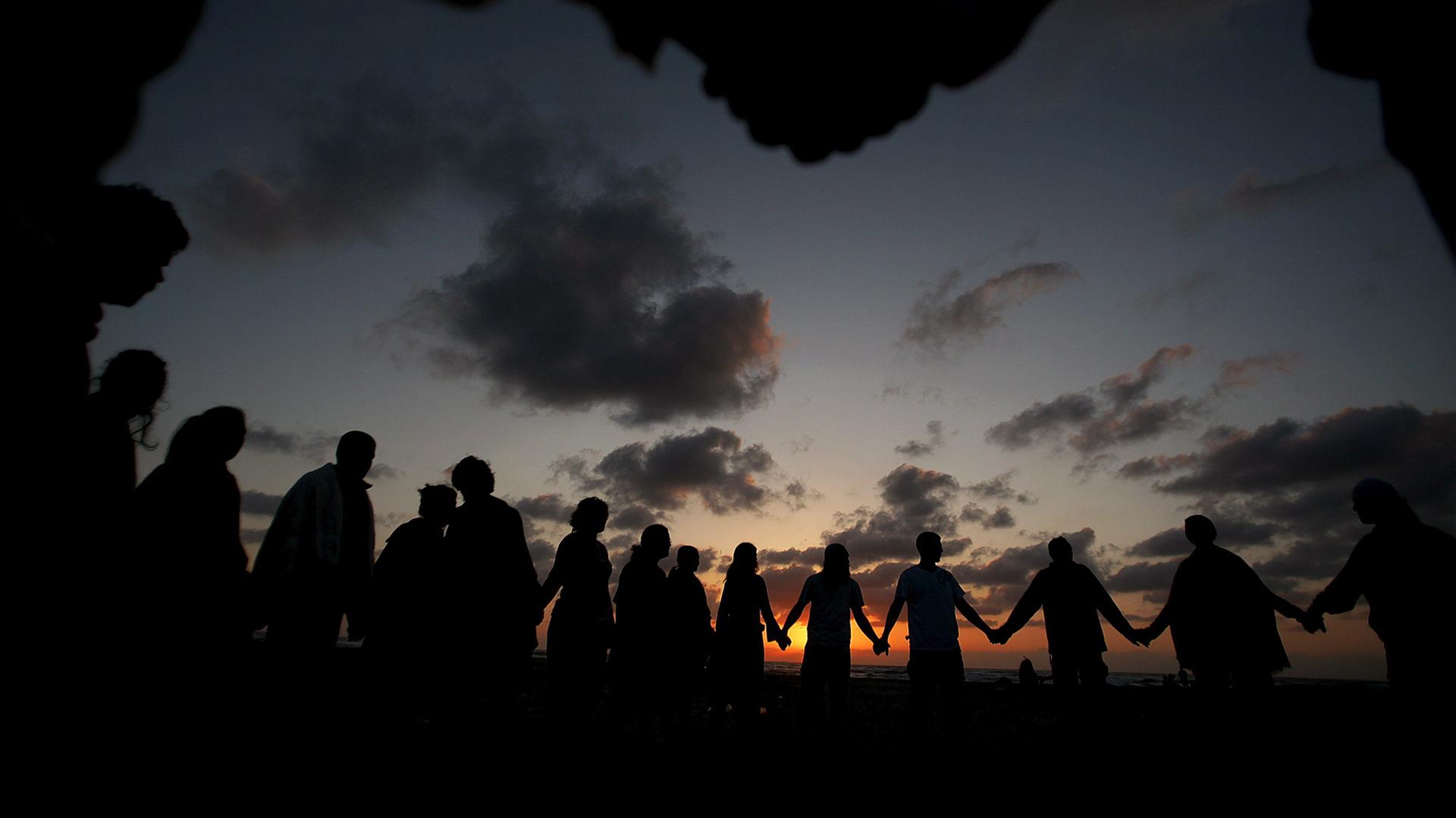 Menschen stehen bei Sonnenuntergang Hand in Hand nebeneinander. | picture-alliance/ dpa