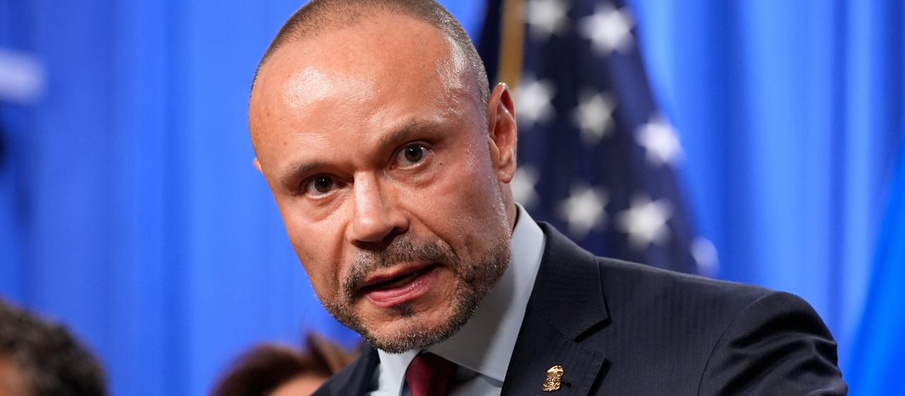 Dan Bongino schaut direkt in die Kamera, hinter ihm verschwommen eine US-Flagge.