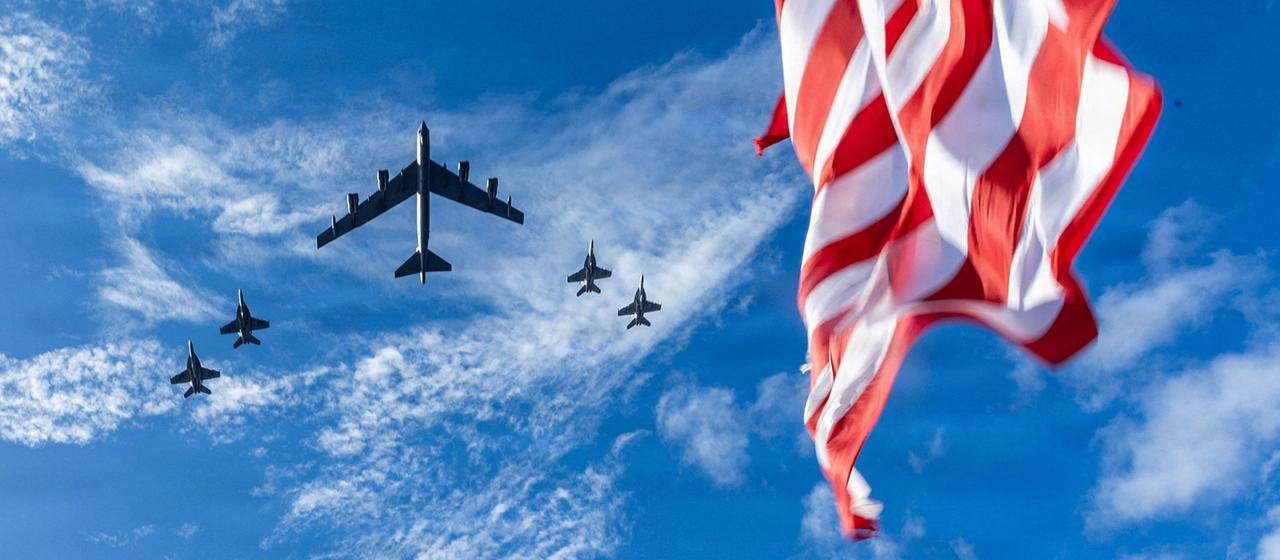 Fünf US-Militärflugzeuge fliegen am Himmel, darunter ein Bomber und vier Kampfjets. Im Vordergrund ist eine wehende amerikanische Flagge zu sehen.