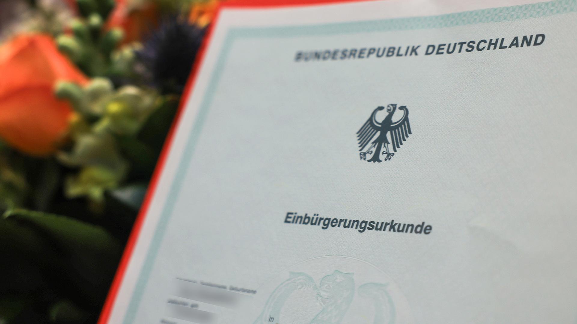Eine Einbürgerungsurkunde wird vor einem Blumenstrauß gehalten. | dpa