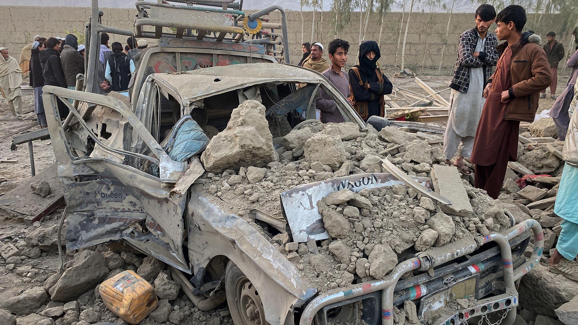 Menschen stehen neben einem von Trümmern überdeckten, zerstörten Fahrzeug. | AP