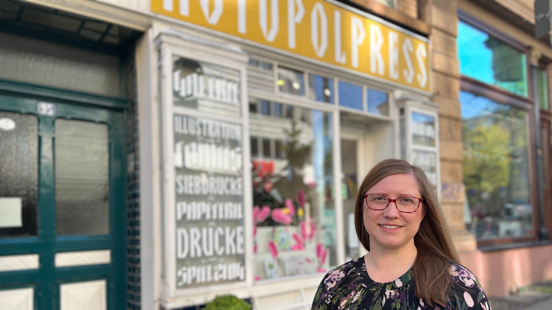 Ein Gründerzeithaus. Davor eine Frau mit langen Haaren, Brille und Kleid. Im Erdgeschoss ein Ladengeschäft mit weißen Fenstern. Im Schaufenster ein Insekt und stilisierte Blumen, ausgeschnitten aus Pappe | Stefanie Küster (hr)