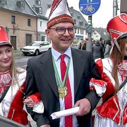 Mann mit Narrenkappe, geleitet von zwei Frauen in Gardekostüm
