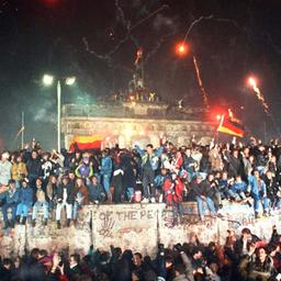 Zahlreiche Menschen sitzen auf und stehen vor der Berliner Mauer, im Hintergrund ist das Brandenburger Tor zu sehen, Pyrotechnik leuchtet in der Luft.