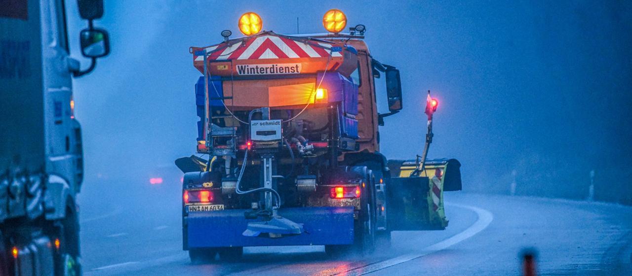 A gritting vehicle drives along a street at dusk.