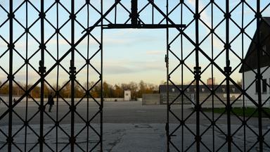 Durch einen Durchgang sieht man durch einen Zaun auf ein offenes Gelände mit Gebäuden.