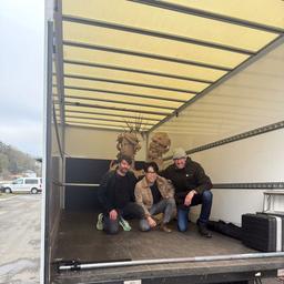 Zu sehen ist die Ladefläche eines kleinen Lastwagens. Darauf sitzen drei Männer. Im Hintergrund sieht man eine Kunst-Skulptur. Die Skulptur aus Pappe hat zwei große Köpfe, einen weiblichen und einen männlichen.