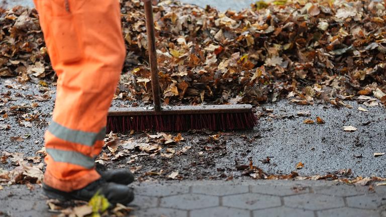 Ein Mitarbeiter der Stadtreinigung beseitigt das Laub mit einem Besen vom Gehweg.
