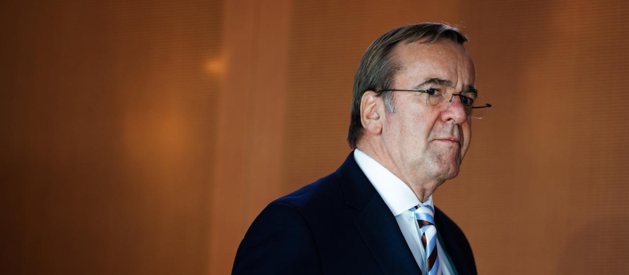 Boris Pistorius geht vor einer braunen Wand entlang.