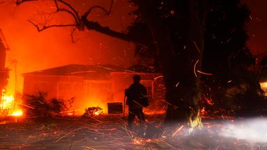 Ein Mensch richtet den Wasserstrahl eines Schlauches auf ein brennendes Haus.