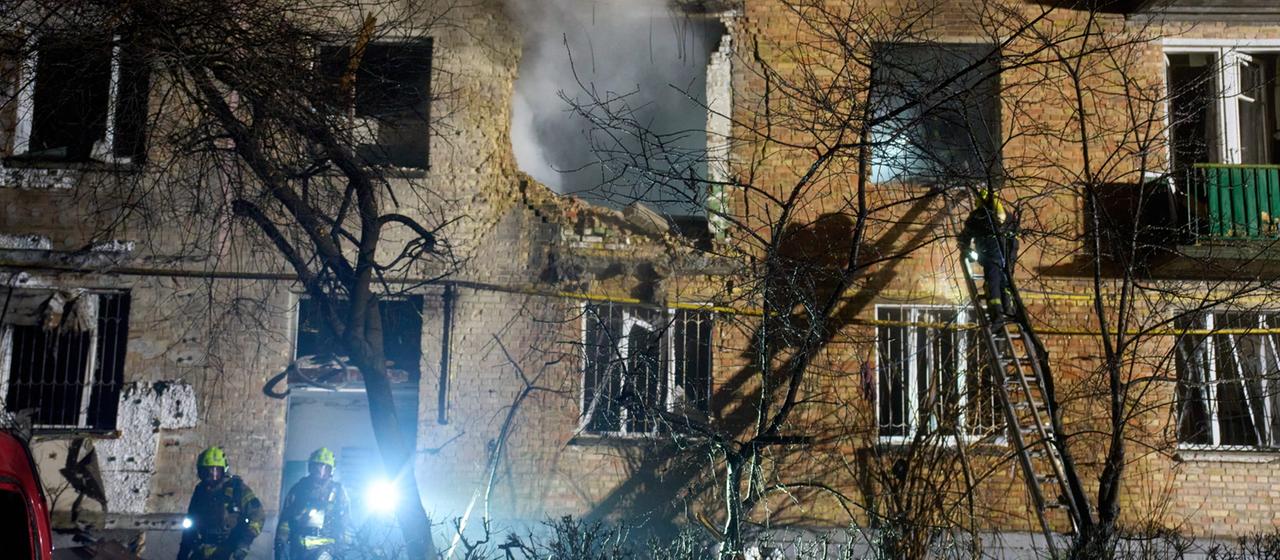 Feuerwehrleute in der Nacht vor einem zerstörten Wohnhaus in Kiew, aus dem Haus dringt Rauch.
