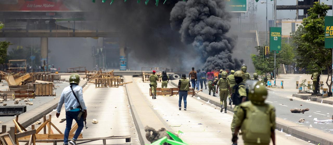 Sicherheitskräfte und Demonstranten laufen in einer Straße in Daressalam, Tansania. Im Hintergrund steigt eine große Rauchwolke auf.
