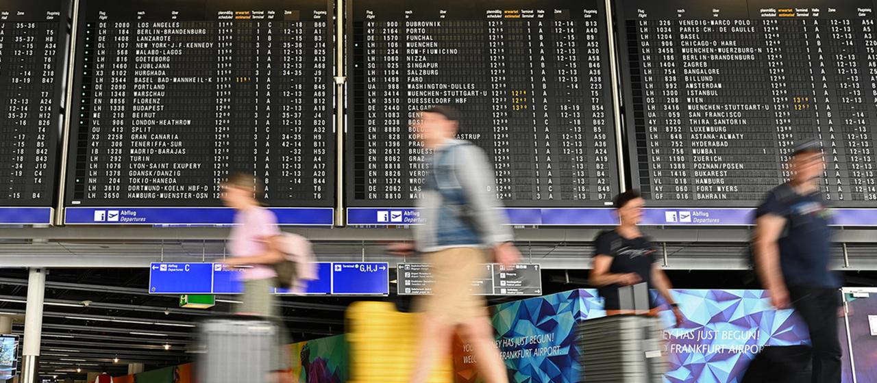 Urlauber laufen im Terminal 1 am Frankfurter Flughafen an einer großen Anzeigetafel vorbei.