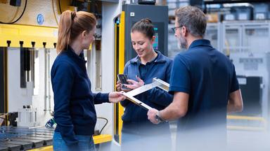 Mitarbeiterinnen und ein Mitarbeiter der Metallindustrie sind während eines Gesprächs am Arbeitsplatz zu sehen.