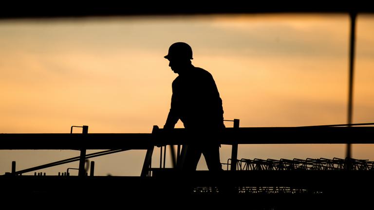 Die Silhouette eines Bauarbeiters zeichnet sich auf einer Baustelle vor dem verfärbten Morgenhimmel ab.