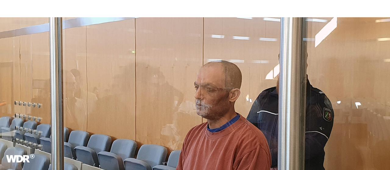 Der Angeklagte steht im Gerichtssaal. Hinter einer Säule steht zur Bewachung ein Wächter.