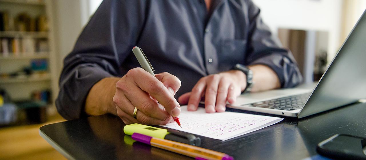 Ein Mann schreibt mit einem Stift auf ein Blatt Papier neben einem Laptop.