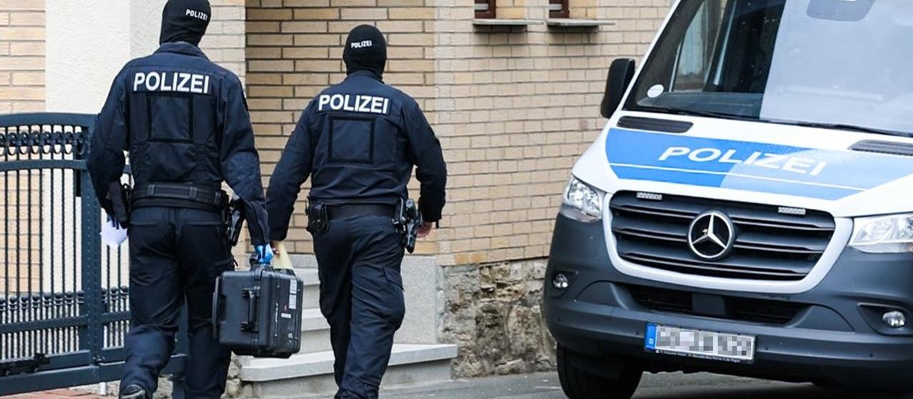Zwei Polizisten gehen zu einem Gebäude, einer trägt einen schwarzen Koffer. Daneben steht ein Polizeiwagen.