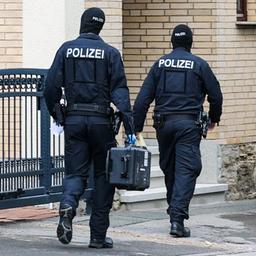 Zwei Polizisten gehen zu einem Gebäude, einer trägt einen schwarzen Koffer. Daneben steht ein Polizeiwagen.