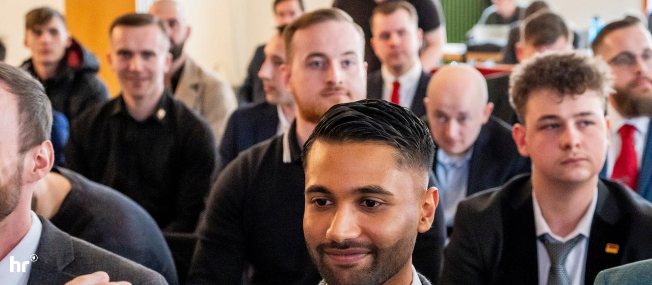 Ein Mann in Anzug (Nafiur Rahman) blickt in eine Handykamera, er ist umgeben von Leuten.