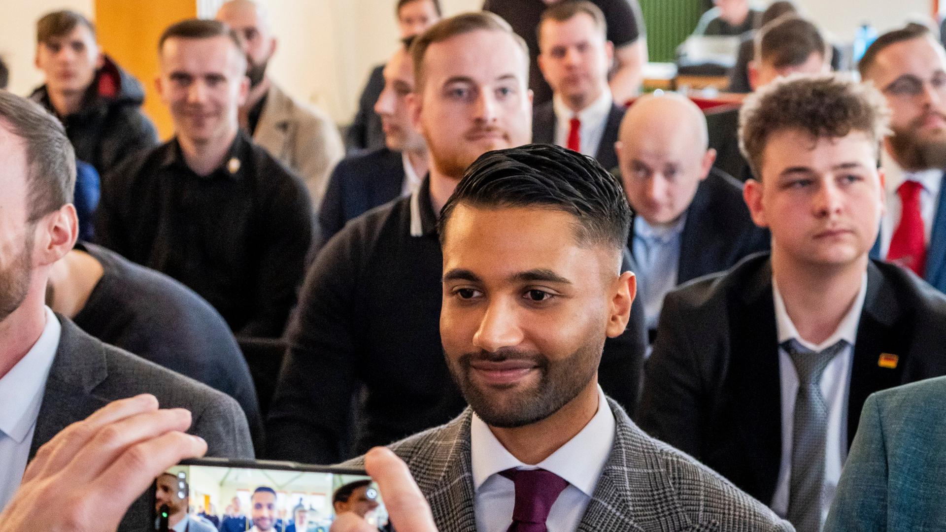 Ein Mann in Anzug (Nafiur Rahman) blickt in eine Handykamera, er ist umgeben von Leuten. | picture alliance/dpa | Andreas Arnold