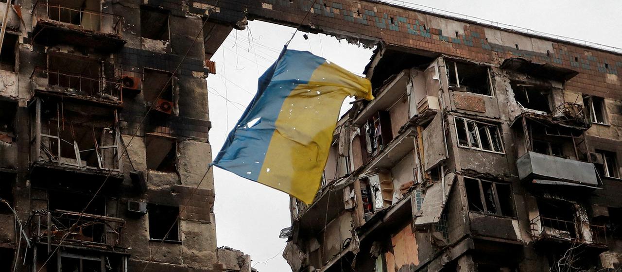 Eine zerrissene ukrainische Flagge hängt an einem Draht vor einem Wohnhaus, das während des russischen Angriffskrieges in der südukrainischen Hafenstadt Mariupol am 14. April 2022 zerstört wurde. | REUTERS Eine zerrissene ukrainische Flagge hängt an einem Draht vor einem Wohnhaus, das während des russischen Angriffskrieges in der südukrainischen Hafenstadt Mariupol am 14. April 2022 zerstört wurde.