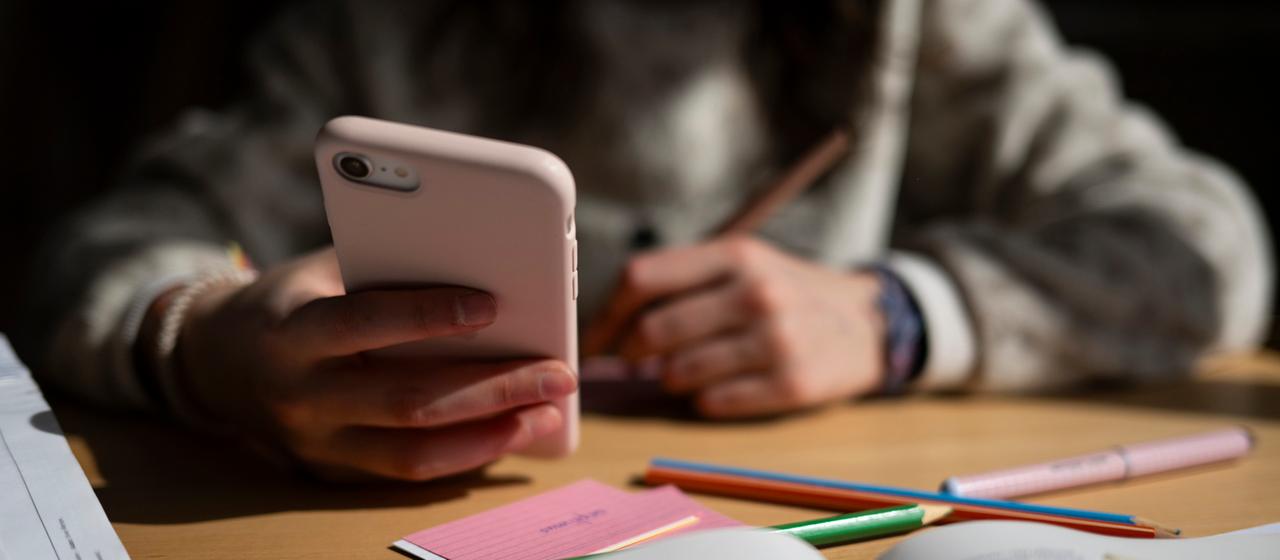 Ein Schulkind sitzt am Schreibtisch und hält ein Smartphone in der Hand.